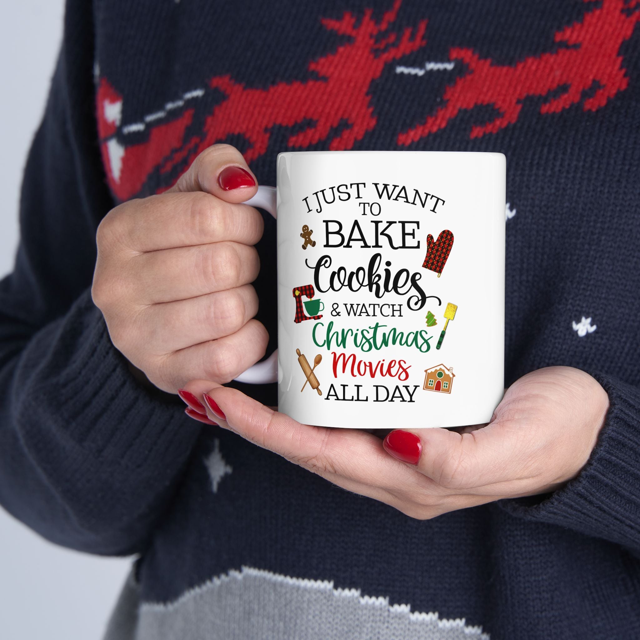 Person holding a white Christmas mug with the quote “I just want to bake cookies and watch Christmas movies all day,” wearing a festive holiday sweater with red reindeer design.