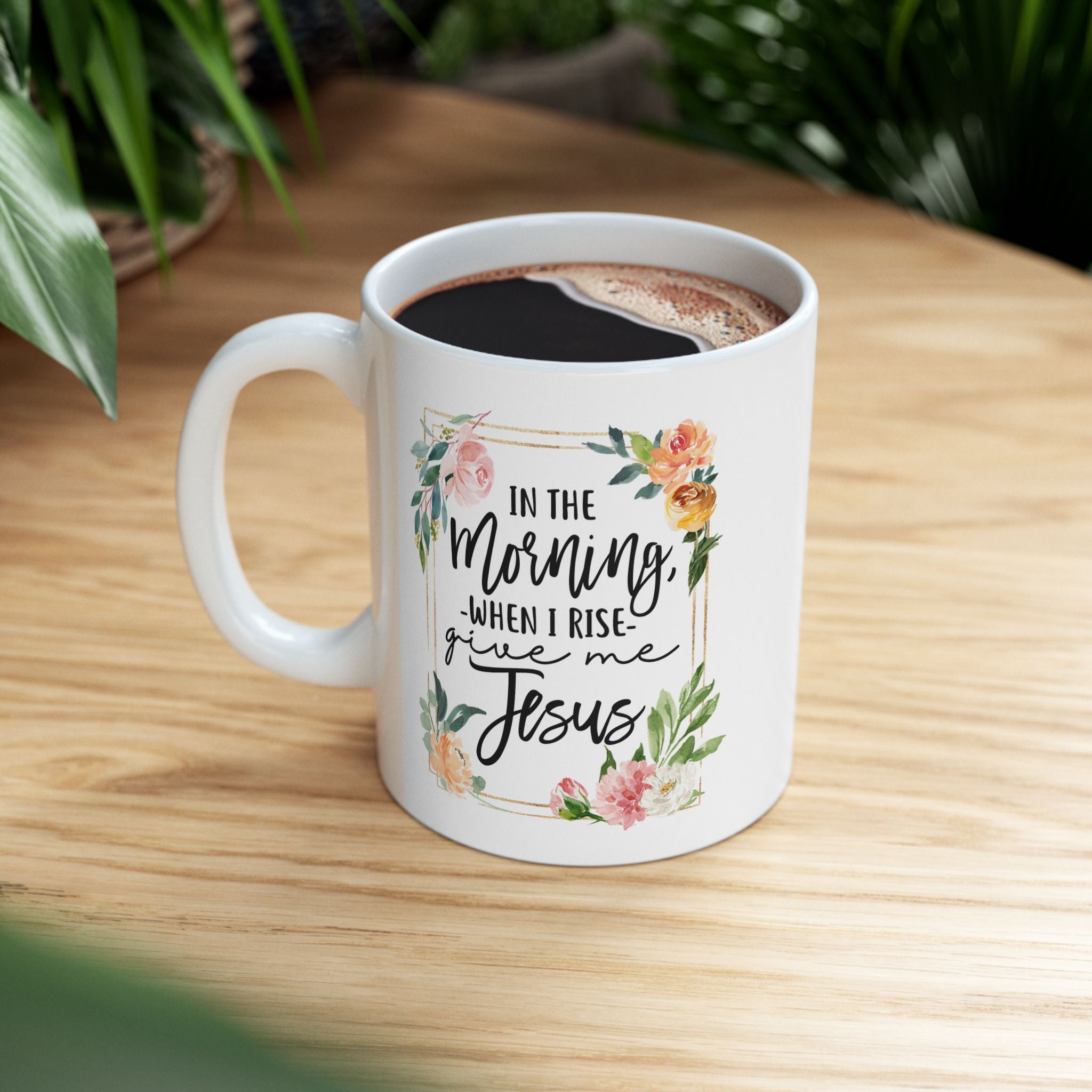 White ceramic coffee mug filled with coffee, featuring floral artwork and the quote “In the Morning When I Rise Give Me Jesus,” sitting on a wooden table with green plants in the background.